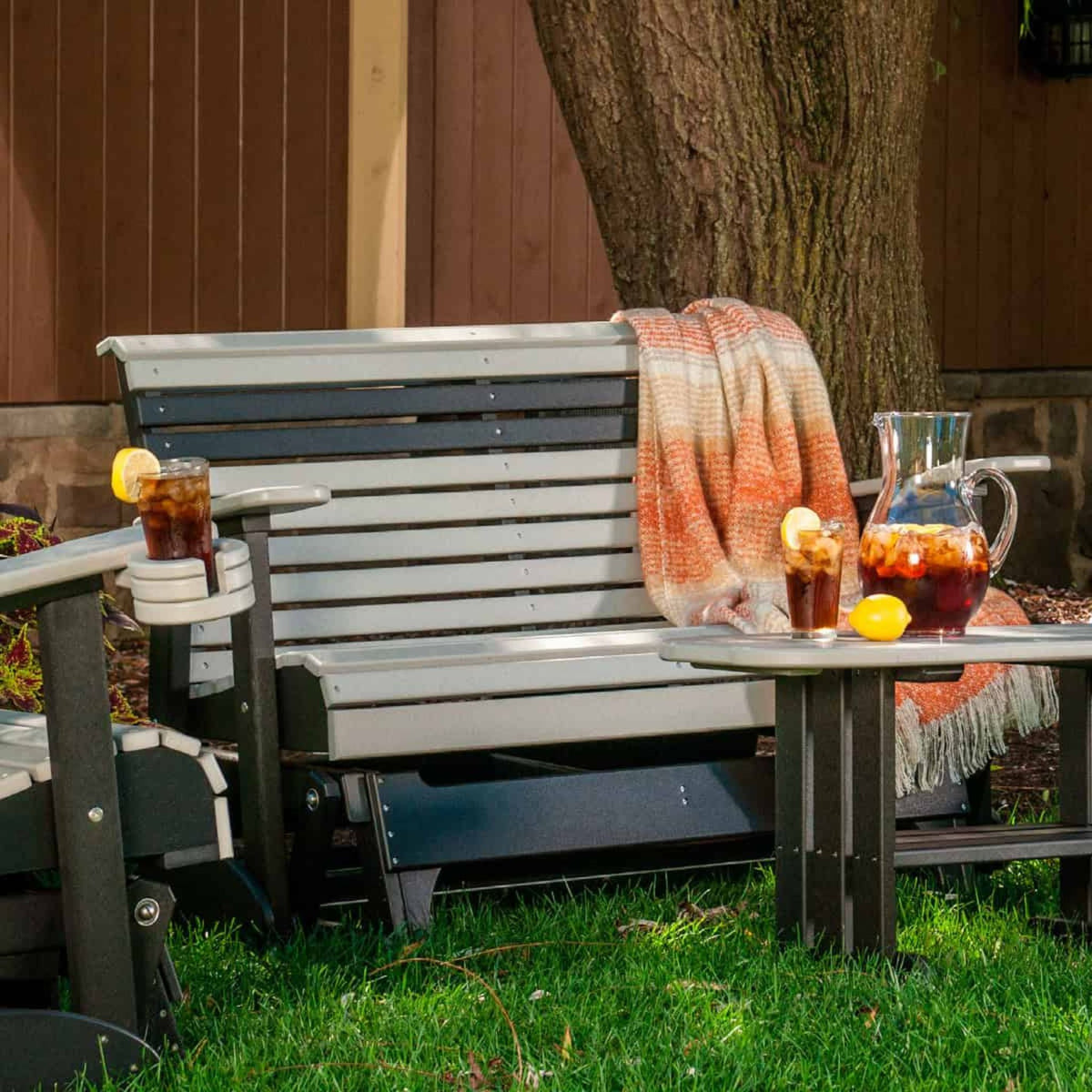 A gray and black outdoor patio bench with a slatted back and armrests, styled with an orange-and-white throw blanket, two iced tea glasses with lemon slices, and a glass pitcher on a small matching side table. The bench is set on grass in front of a wooden fence and tree trunk.