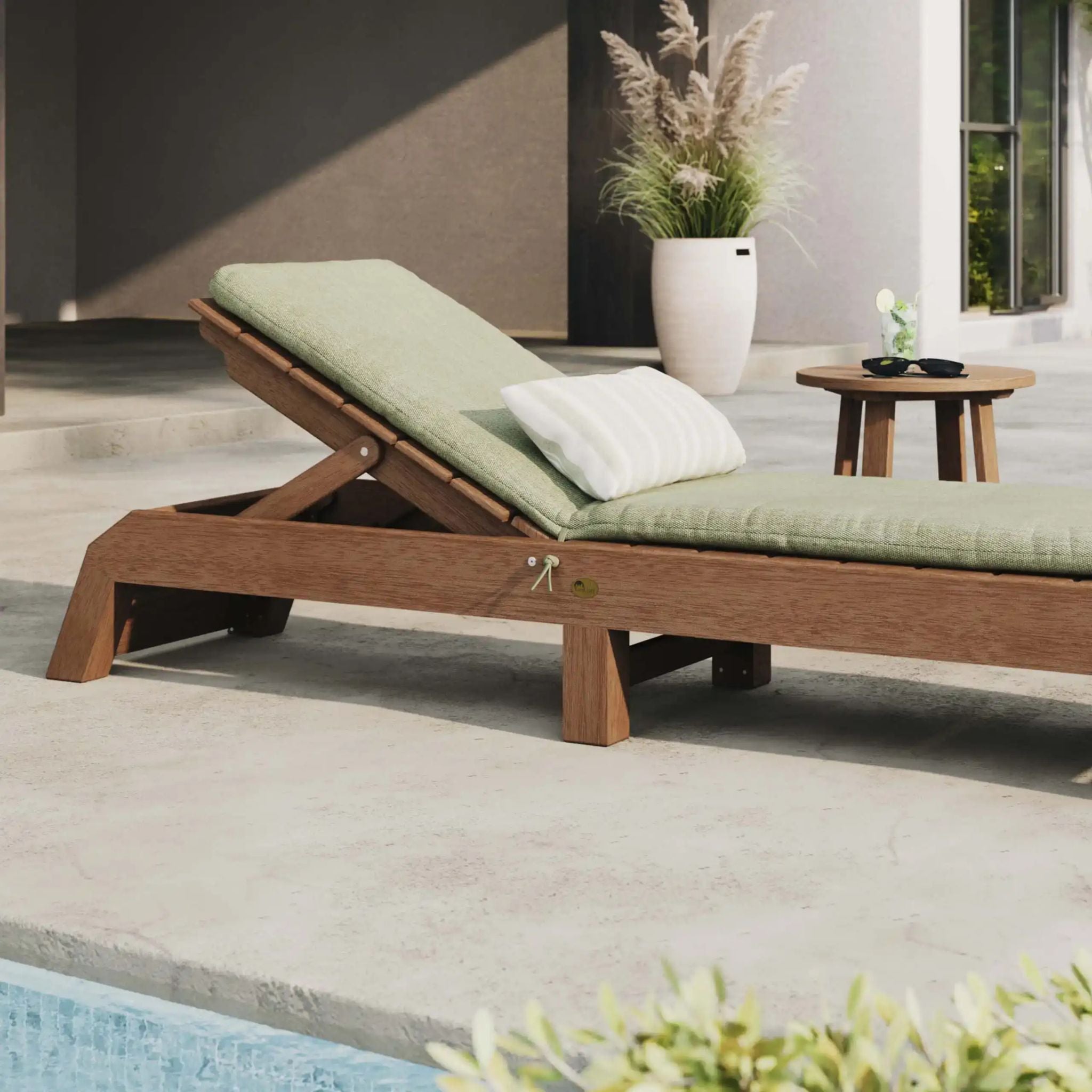 A wooden outdoor chaise lounge with light green cushions and a white pillow, positioned next to a pool on a patio with a small side table and potted plants in the background.