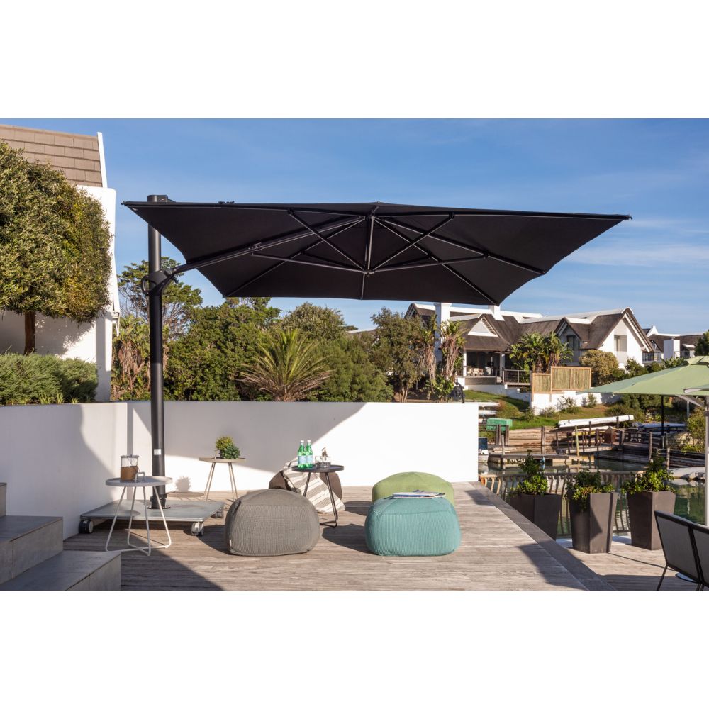 A large black rectangular patio umbrella with a black pole is shown on a wooden outdoor terrace with lounge chairs, a small table, and residential houses in the background under a blue sky.