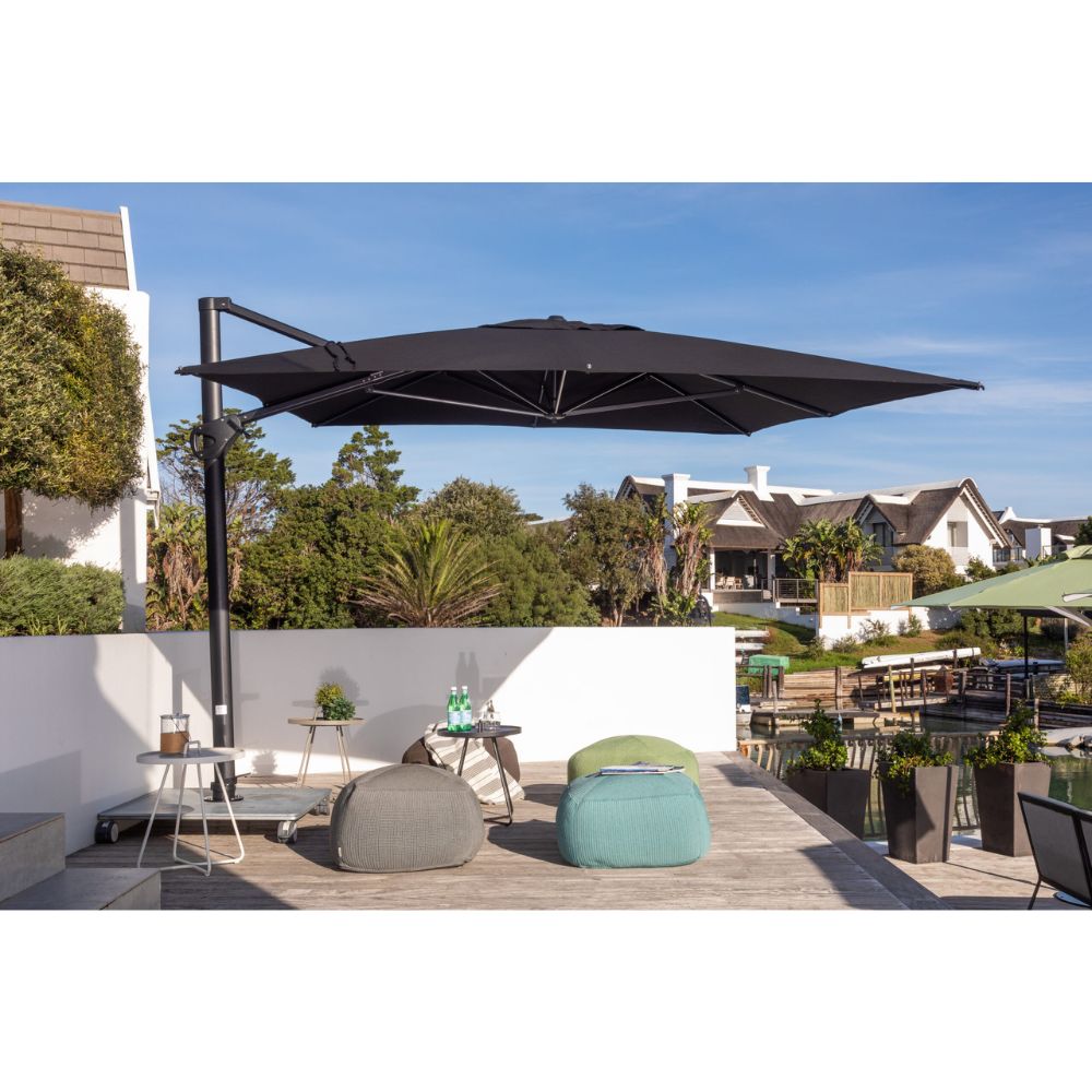 A large black cantilever patio umbrella shading an outdoor deck with lounge chairs, poufs, and a small table, set against a residential background with a white wall and blue sky.