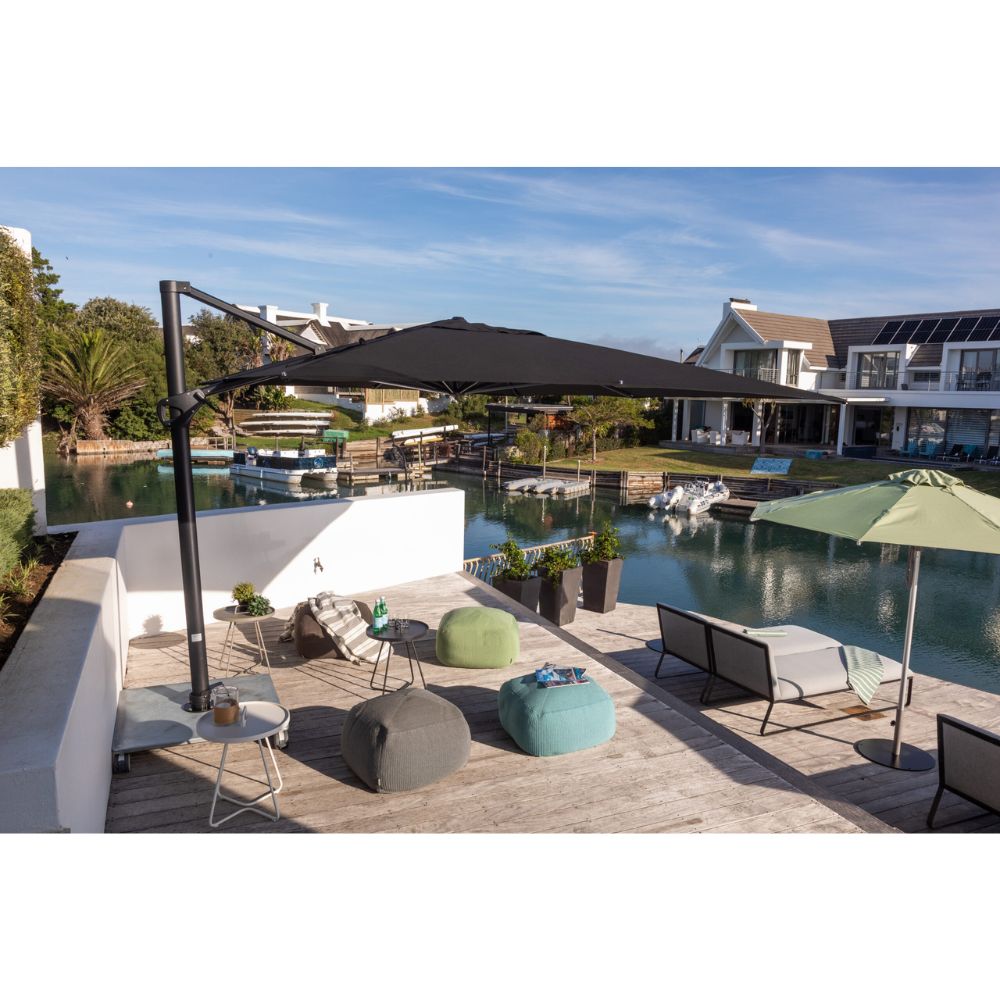 Outdoor patio scene with a large black cantilever umbrella over lounge chairs and bean bags, next to a green patio umbrella by the water, with houses and boats in the background.