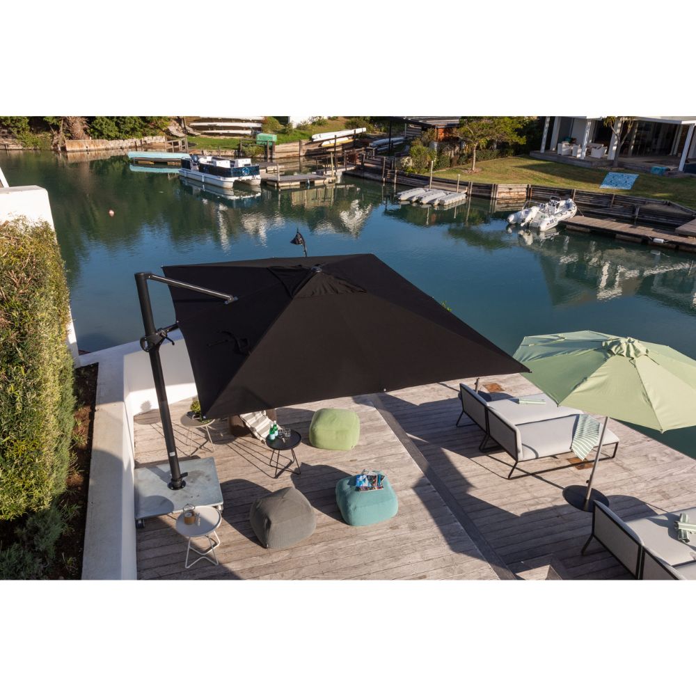 A large black cantilever patio umbrella shades an outdoor deck area with lounge chairs, bean bags, and a light green umbrella, overlooking a canal with boats in the background.