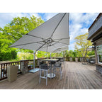 A large square patio umbrella with a light gray canopy and silver metal frame, shading a bar-height outdoor dining table and chairs on a wooden deck. The scene is set on a residential deck with trees and sky in the background.