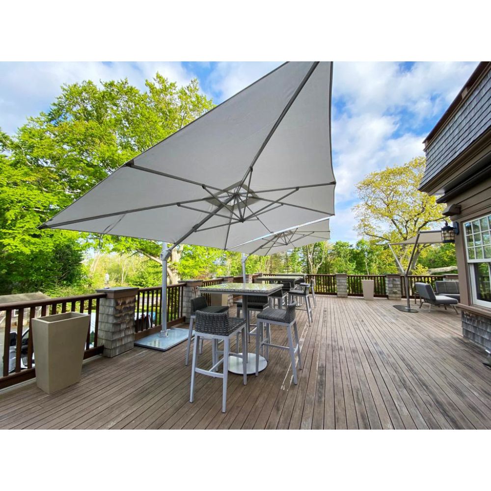 A large square patio umbrella with a light gray canopy and silver metal frame, shading a bar-height outdoor dining table and chairs on a wooden deck. The scene is set on a residential deck with trees and sky in the background.