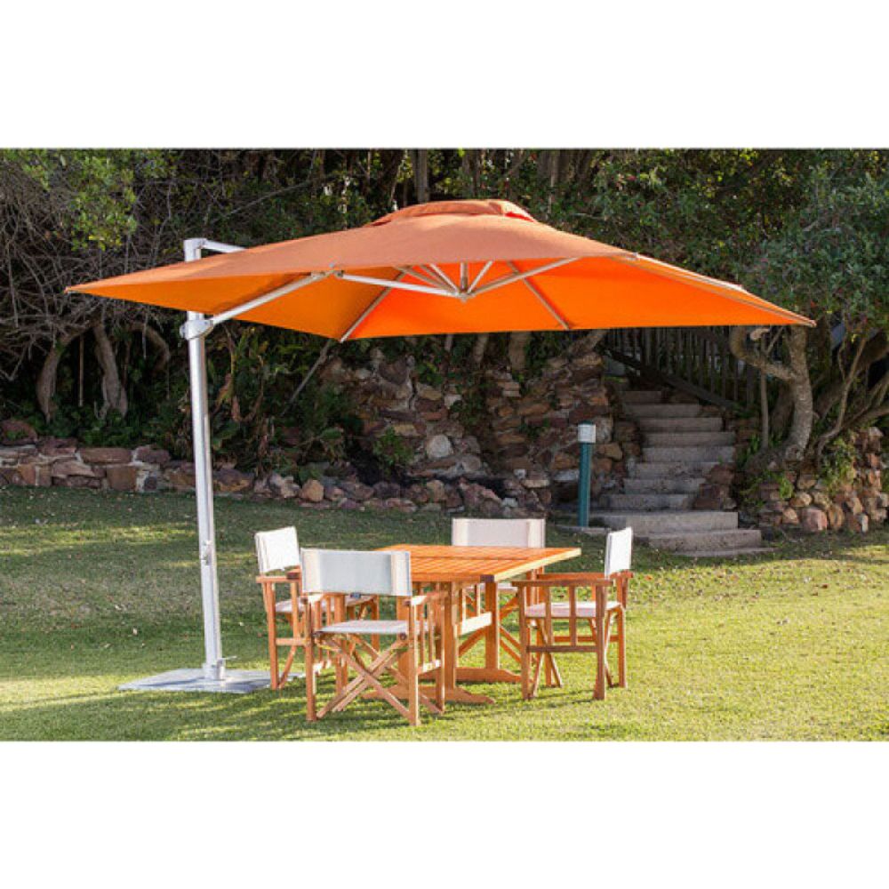 An outdoor patio umbrella with a bright orange canopy and a white pole, shading a wooden dining table and four chairs on a grassy lawn with trees and stone steps in the background.
