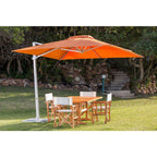 An outdoor patio umbrella with a bright orange canopy and a white pole, shading a wooden dining table and four chairs on a grassy lawn with trees and stone steps in the background.