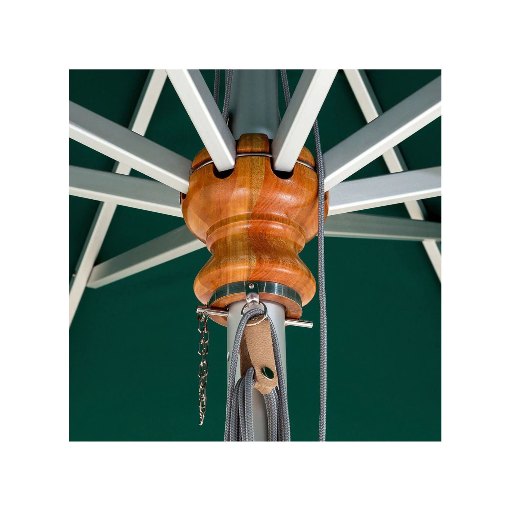 Close-up of a patio umbrella canopy and central wooden finial, showing white support ribs, a warm brown wood top, metal hardware, and hanging cords against a dark green background.