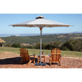 A large white patio umbrella with a silver metal pole stands over two wooden Adirondack chairs and a small side table on a brick patio, with a scenic outdoor landscape in the background under a blue sky.
