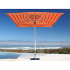 An orange and red striped patio umbrella with a silver metal pole, positioned next to a swimming pool with a beach and ocean view in the background.