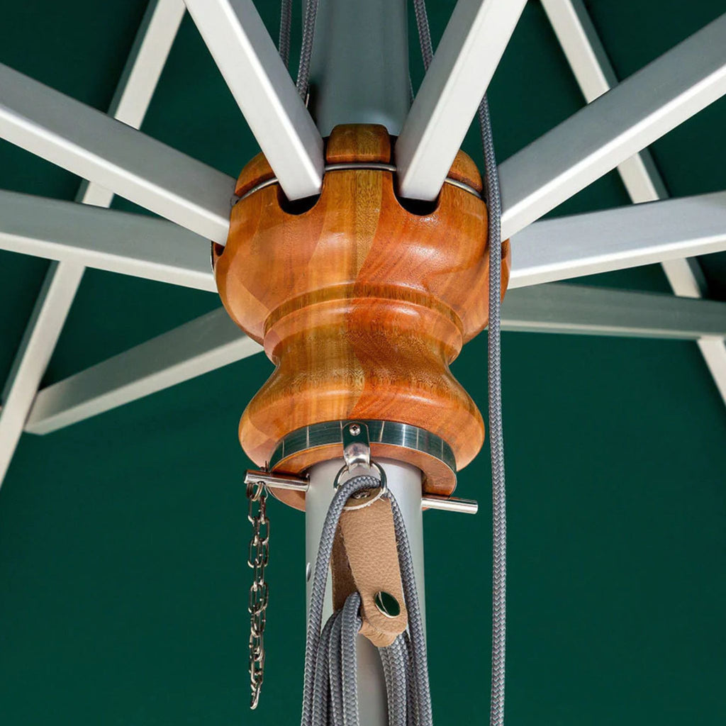 Close-up of a wooden umbrella pull ring and canopy attachment hardware on a green patio umbrella, showing the warm brown wood grain, silver metal bands, and gray cords.