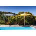 Two outdoor patio umbrellas, one white and one yellow, positioned on a wooden deck next to a swimming pool with a lush green garden and blue sky in the background.
