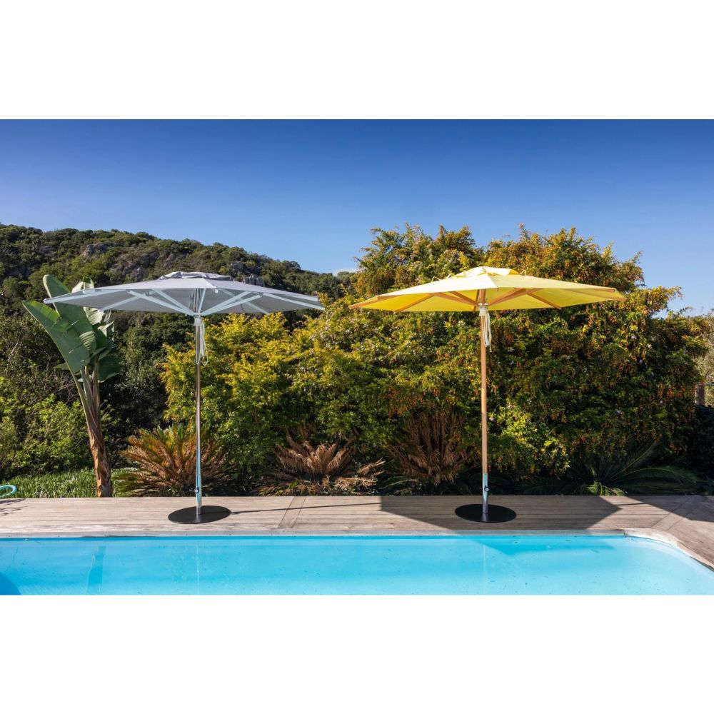 Two outdoor patio umbrellas, one white and one yellow, positioned on a wooden deck next to a swimming pool with a lush green garden and blue sky in the background.