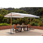 A large outdoor patio canopy with a white canopy fabric and orange-brown support poles, shading a rectangular dining table and four wooden chairs on a concrete patio. The background features a lush green garden and rocky hillside.