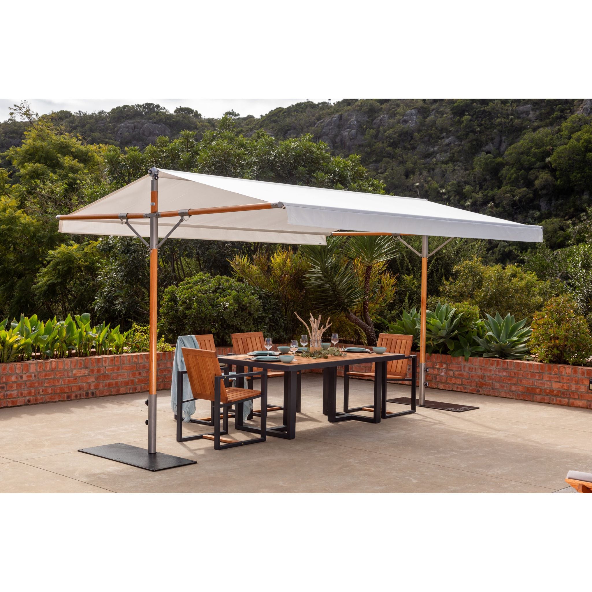 A large outdoor patio canopy with a white canopy fabric and orange-brown support poles, shading a rectangular dining table and four wooden chairs on a concrete patio. The background features a lush green garden and rocky hillside.
