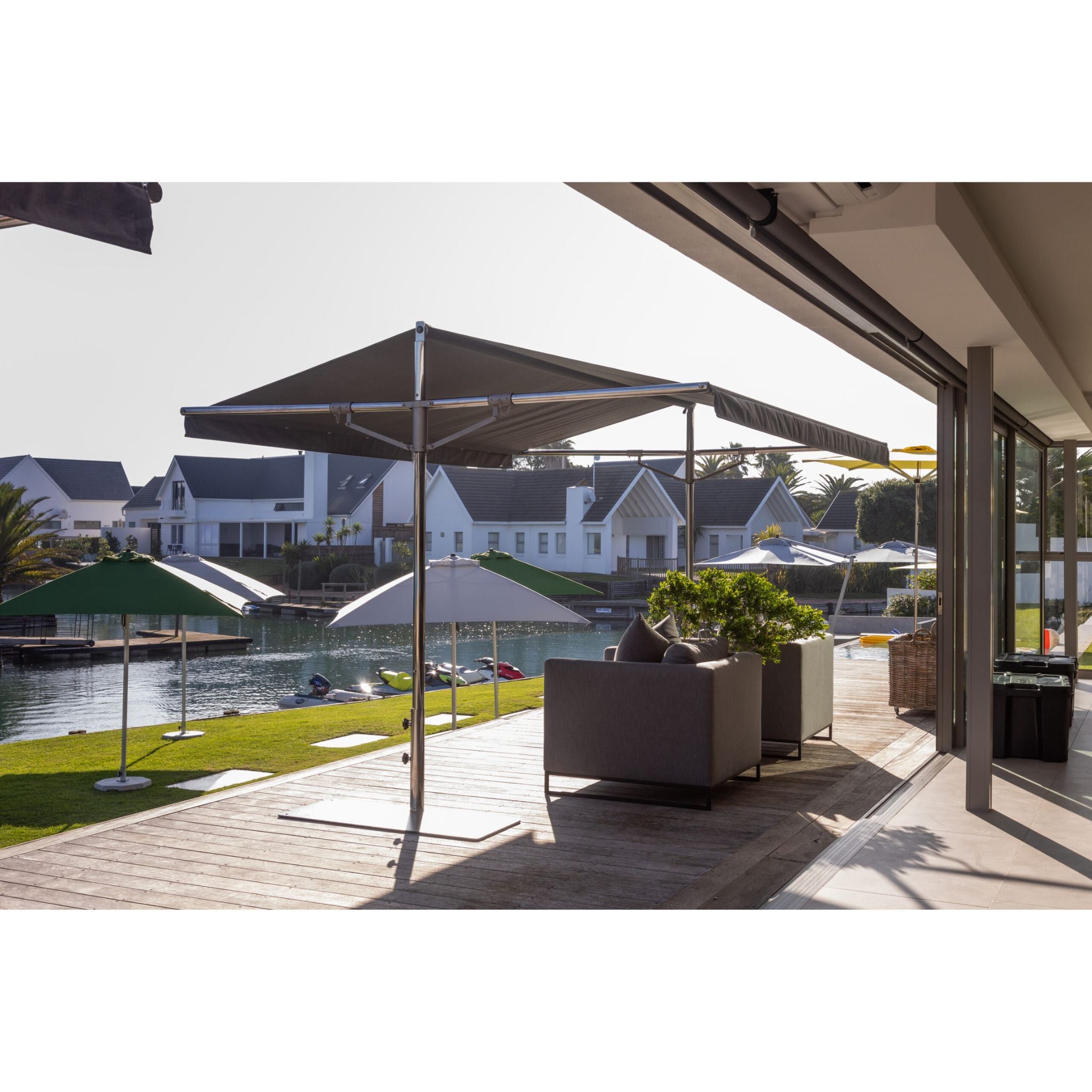 A modern patio umbrella with a dark gray fabric canopy and a chrome metal frame, set up on a wooden deck next to a house with a lake view.