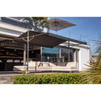 A modern outdoor patio setup featuring a large dark gray cantilevered patio umbrella over a beige sectional sofa, with a glass-walled contemporary house in the background.