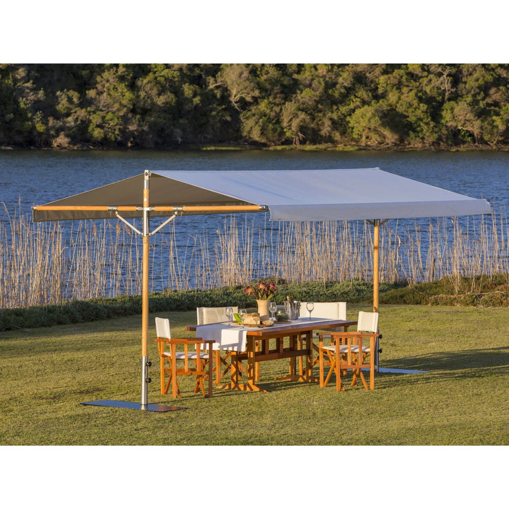 A large outdoor patio umbrella with a light gray canopy and wooden frame is set up on a grassy lawn next to a dining table and chairs, with a lake and trees in the background.