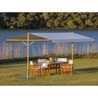 A large outdoor patio umbrella with a light gray canopy and wooden frame is set up on a grassy lawn next to a dining table and chairs, with a lake and trees in the background.