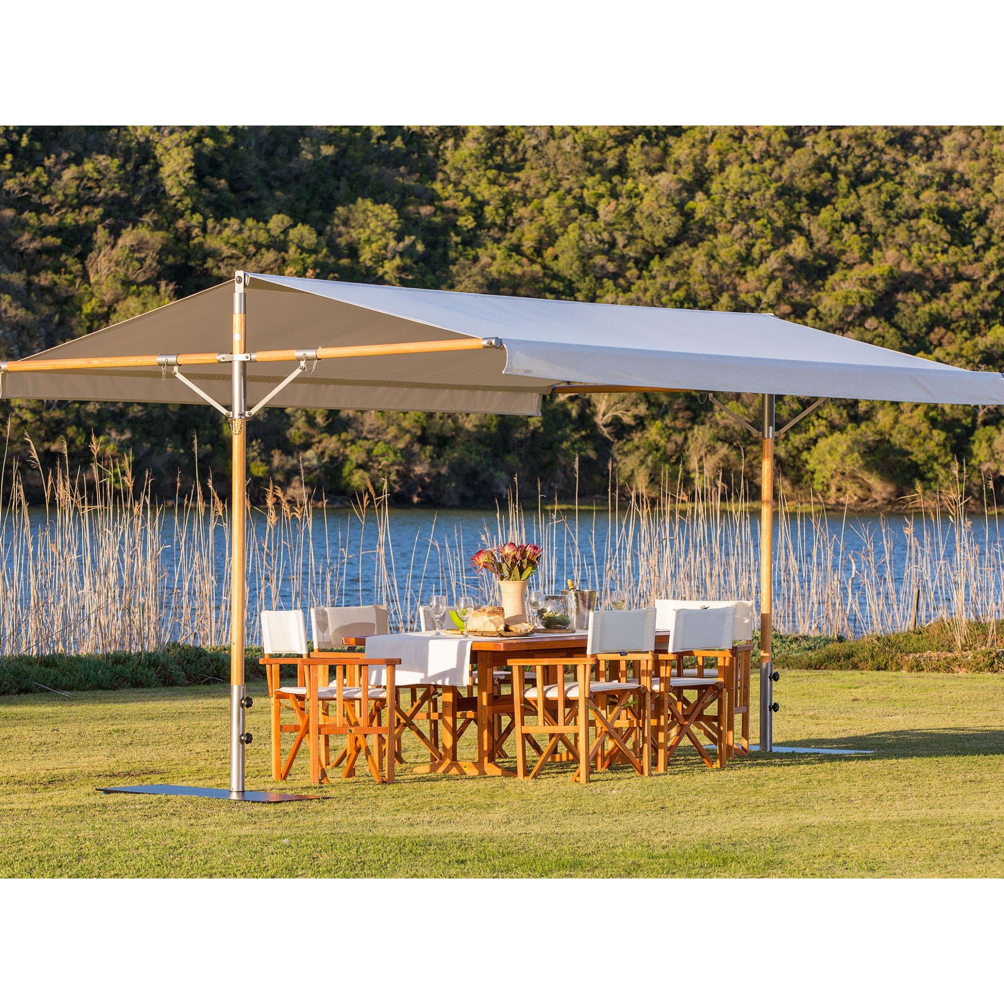 A large beige patio canopy with a dining table and four white-cushioned wooden chairs underneath, set on a green lawn with a lake and forest in the background.