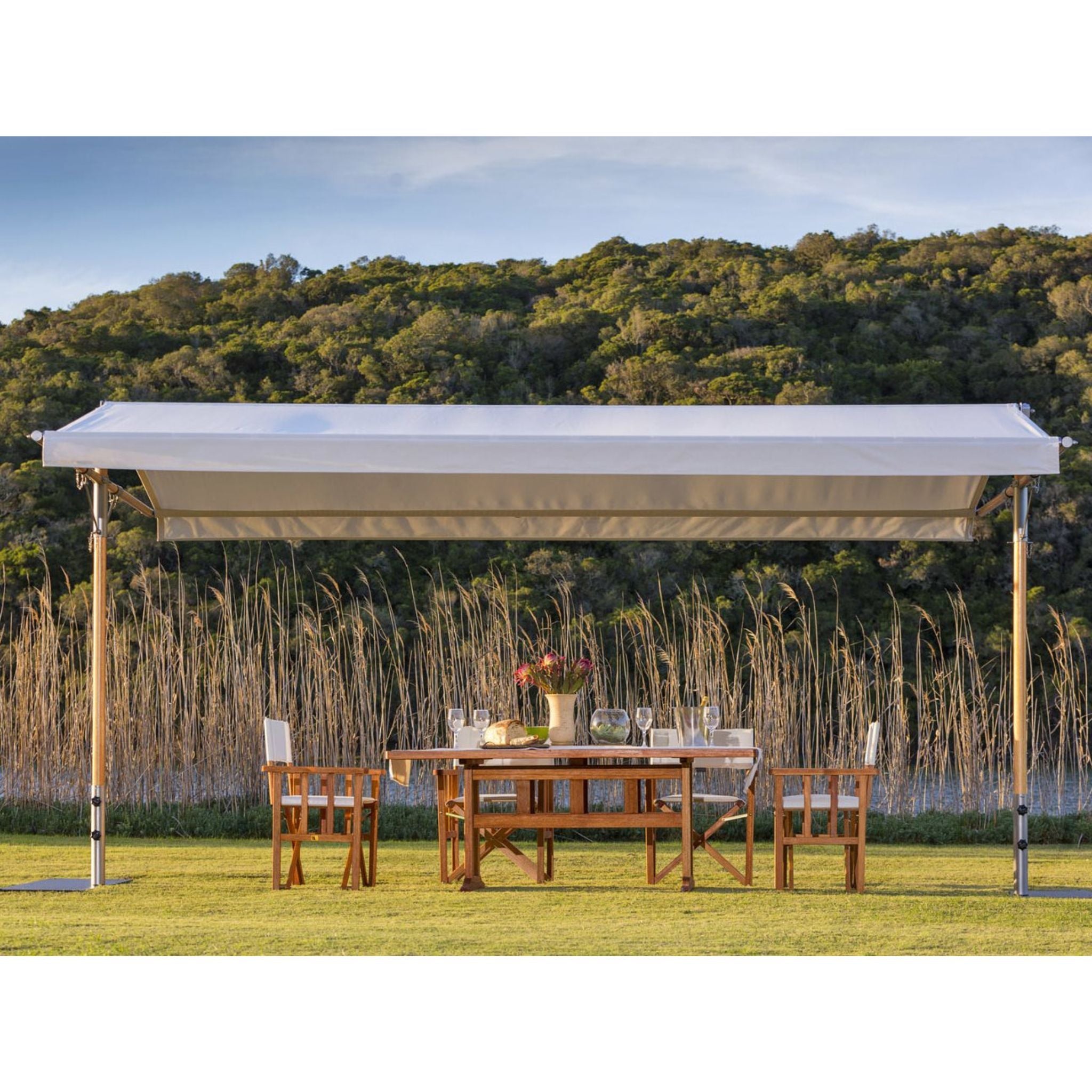 A large outdoor patio canopy with a white fabric roof and light-colored wooden posts, set up on a green lawn with a wooden dining table and chairs underneath. Tall reeds and a forested hill are visible in the background.